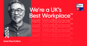 Smiling gentleman with glasses and grey hair featured against a vibrant red background with the repeated text "Best Workplace" and the statement "We’re a UK’s Best Workplaces 2024" alongside the Great Place To Work logo for small organizations.