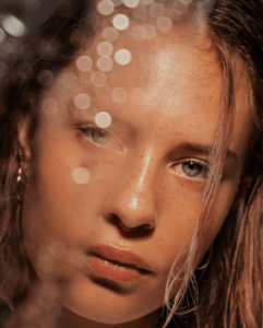 Close-up portrait of a young woman with a nose ring and freckles, partially obscured by soft bokeh highlights, with wet-look hair and warm lighting