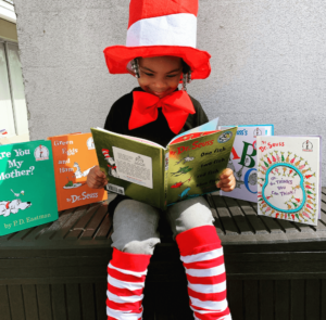 A young child dressed in a red and white striped hat and bow tie reading a Dr Seuss book while seated, surrounded by other Dr Seuss titles