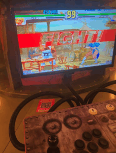 A vintage arcade game cabinet showing a "Street Fighter" match between Ken and Chun-Li, with a game controller in the foreground.